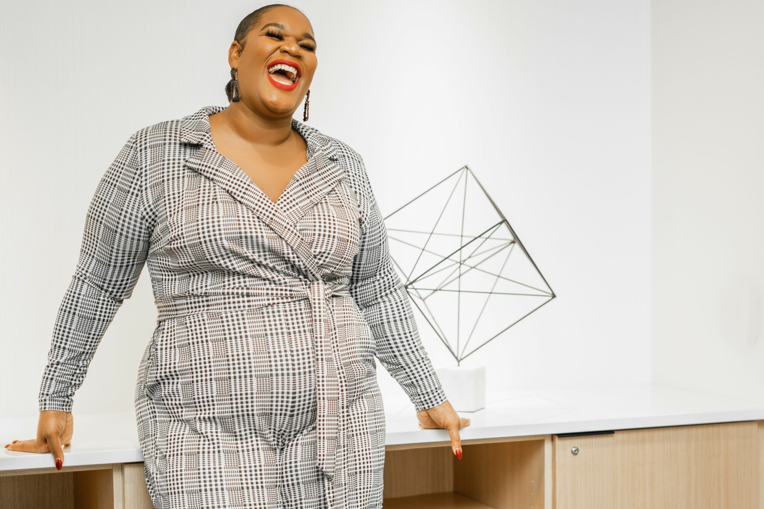 A smiling woman with dark hair and earrings stands behind a table, with a geometric sculpture in the background.