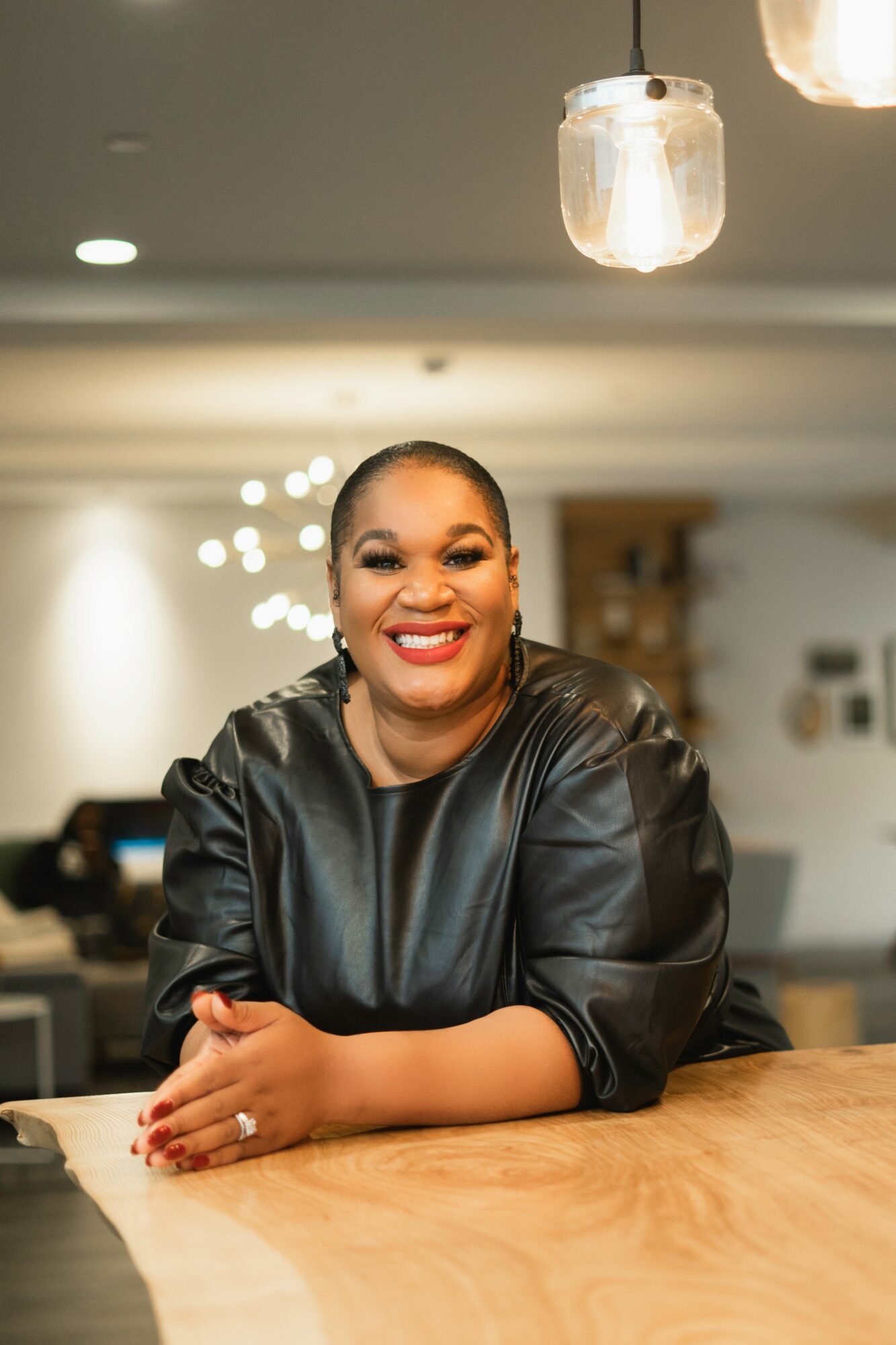 Smiling woman with short hair and makeup, wearing a black leather top, sitting at a wooden table in a modern indoor setting.