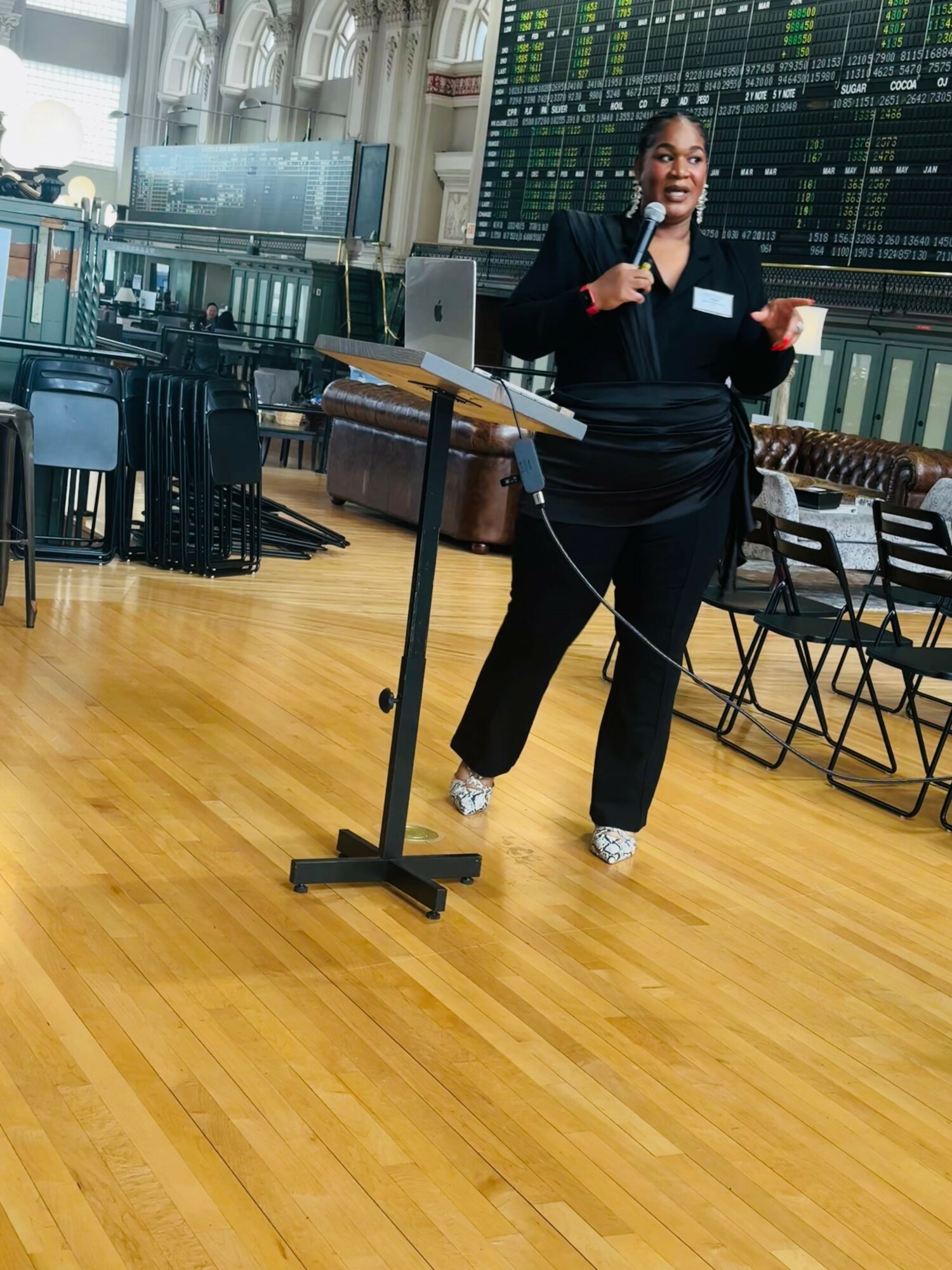 Woman speaking into microphone at a financial trading floor with large screens and wooden floor.