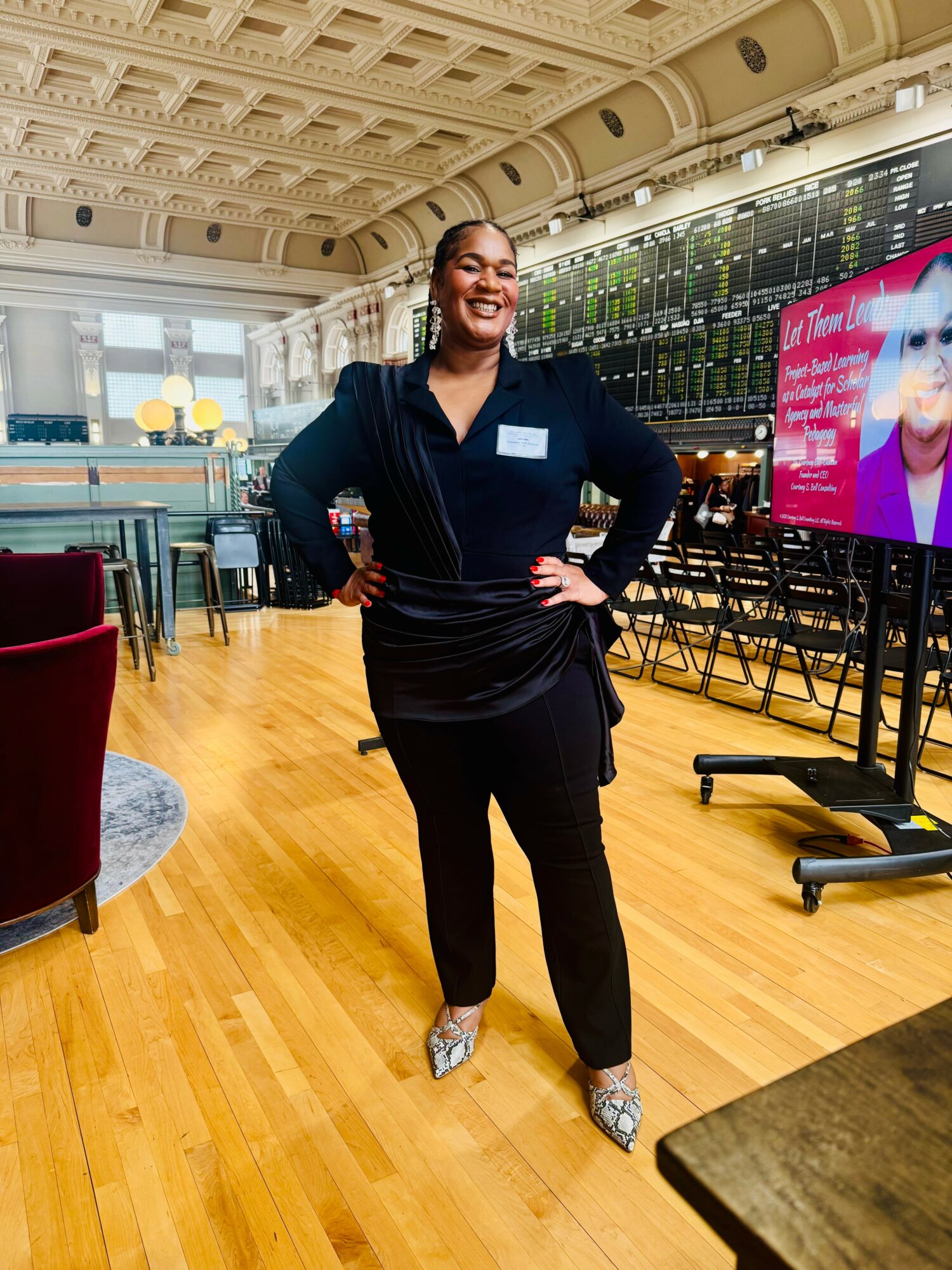 Smiling woman in black outfit standing in a spacious room with wooden floors and large screens, hands on hips.