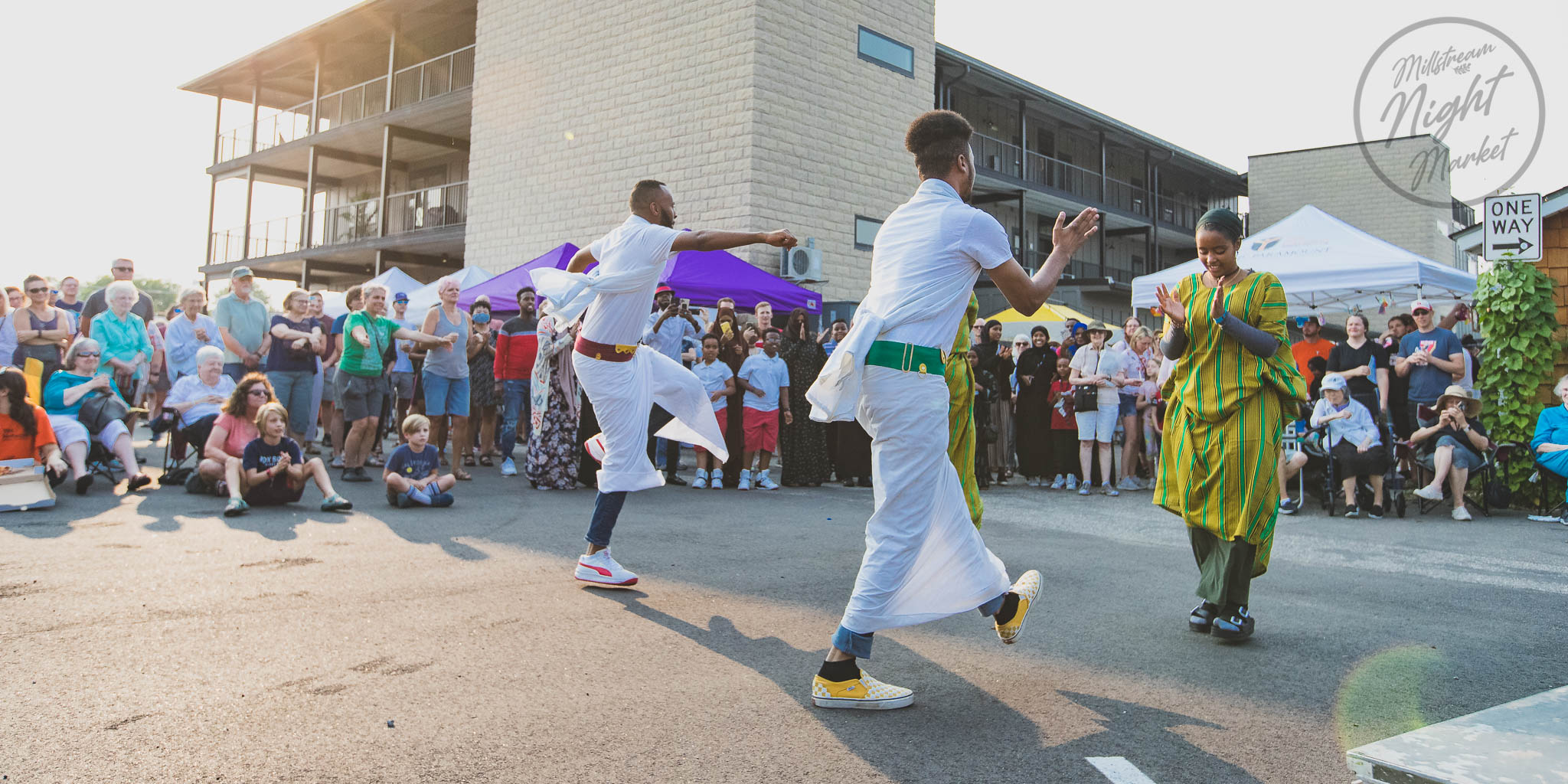 People gather outdoors watching performers dance, with a building and tents in the background.