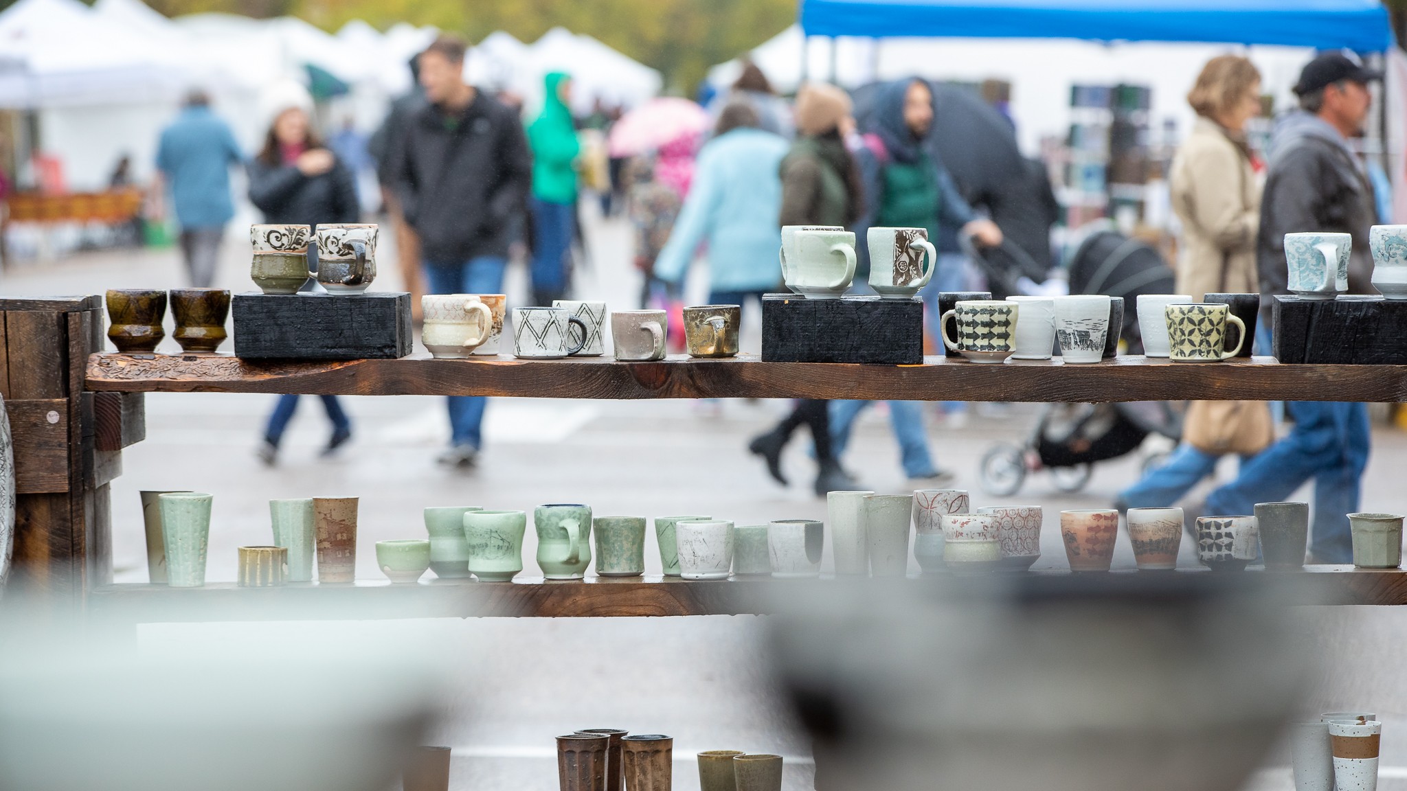 Display of various cups and bowls on a wooden shelf at an outdoor market, with people walking in the background.