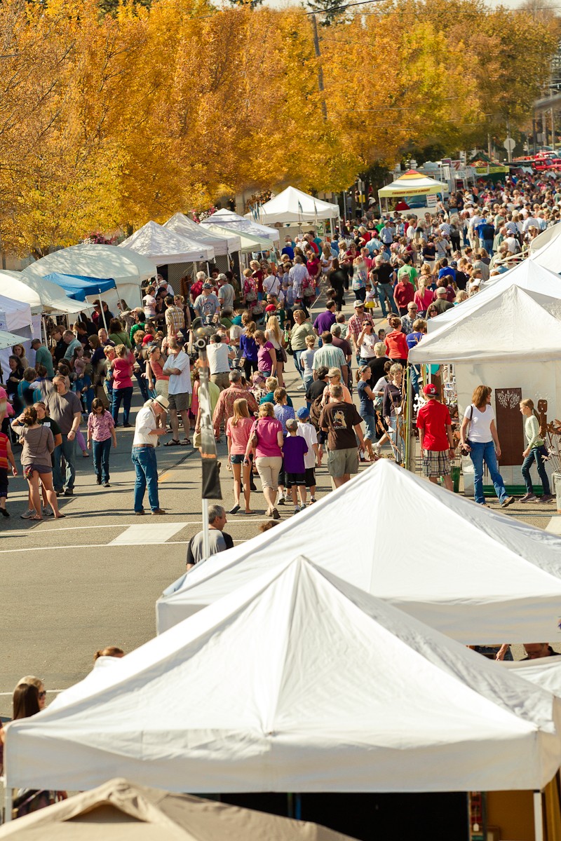 Crowd of people at outdoor market with white tents and autumn trees in background.