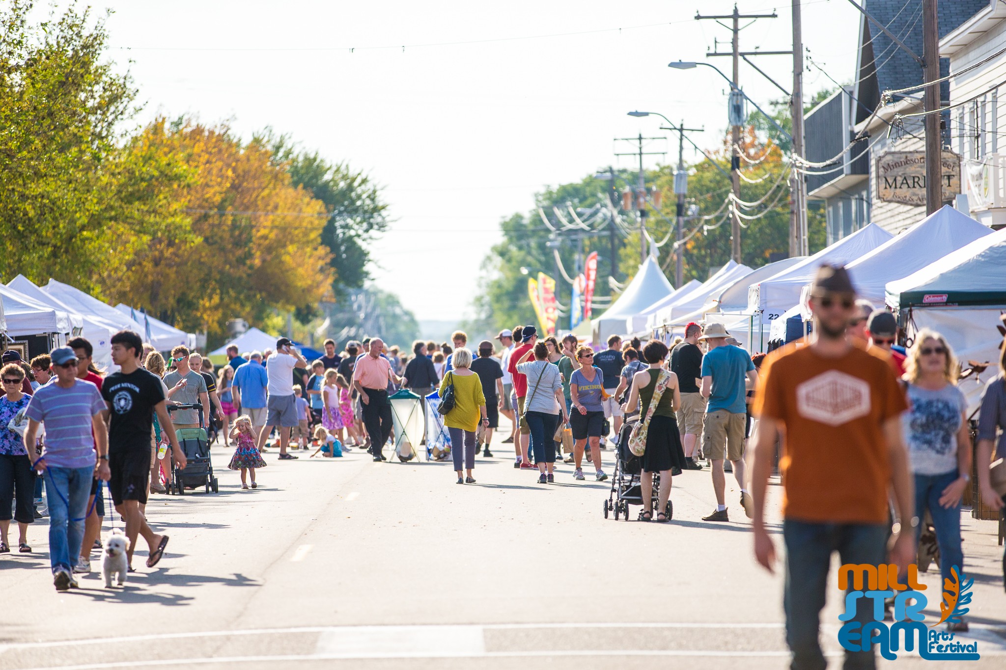 Crowd of people walking along a street with tents on both sides during daytime, trees in the background.