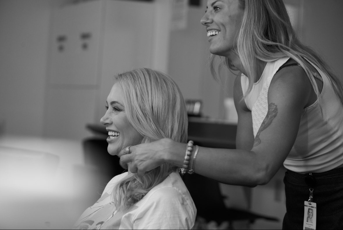Two women smiling, one sitting and the other standing, in a room with cabinets in the background.