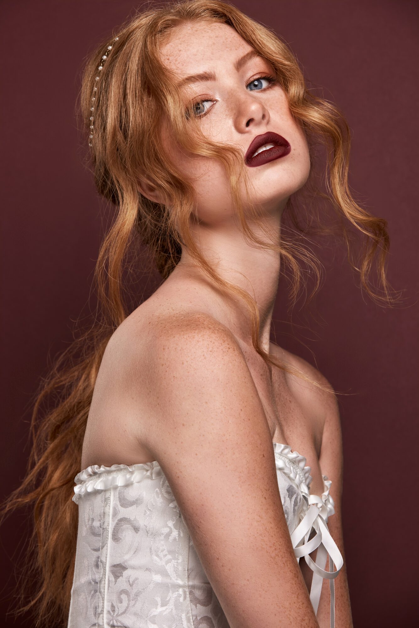 Portrait of a woman with long red hair, dark lipstick, and a white strapless dress, looking at the camera.