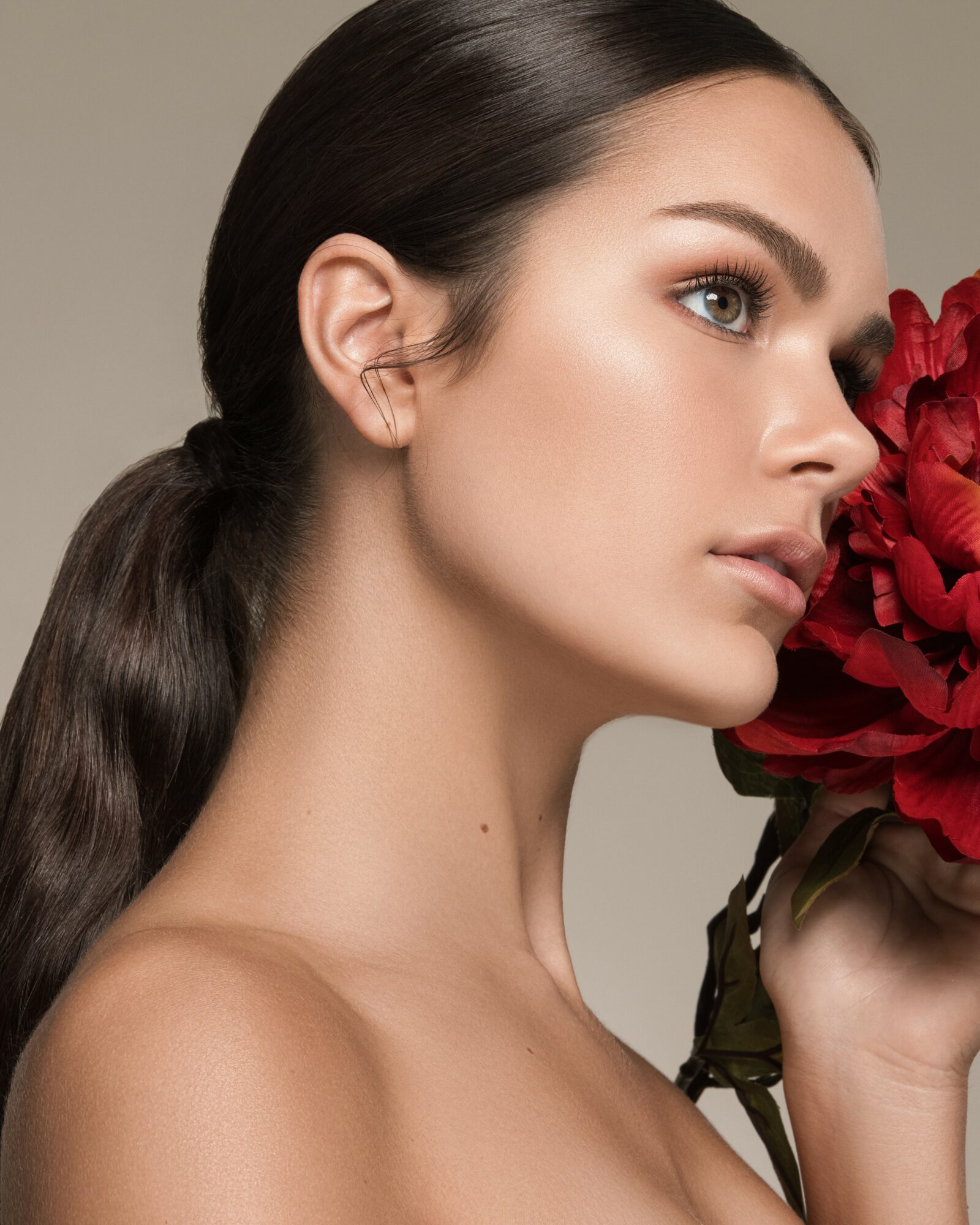 Side profile of a woman with dark hair in a ponytail holding a red flower near her face, neutral background.