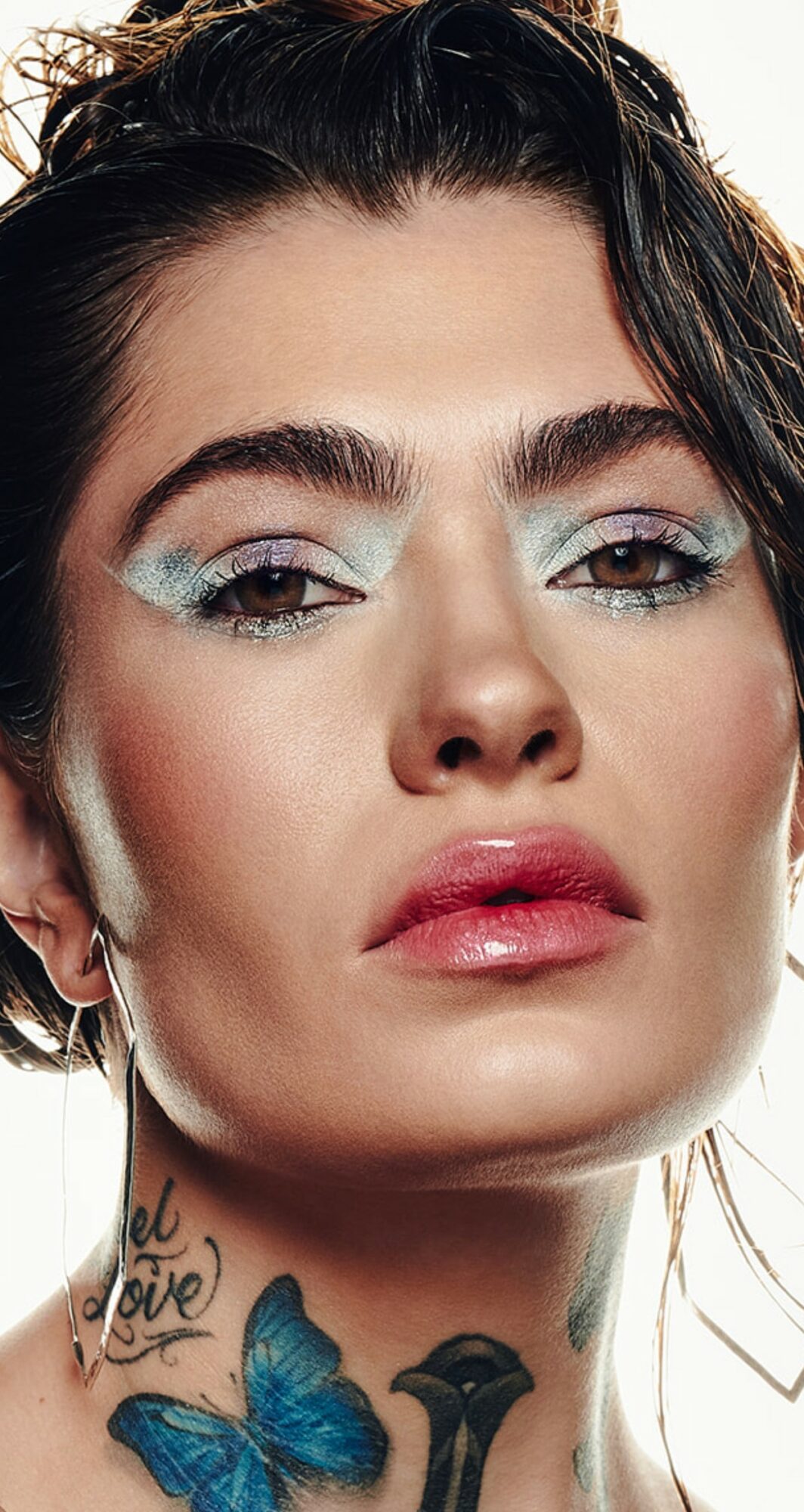 Close-up of a woman with makeup, dark hair, and a tattoo on her neck, looking directly at the camera.