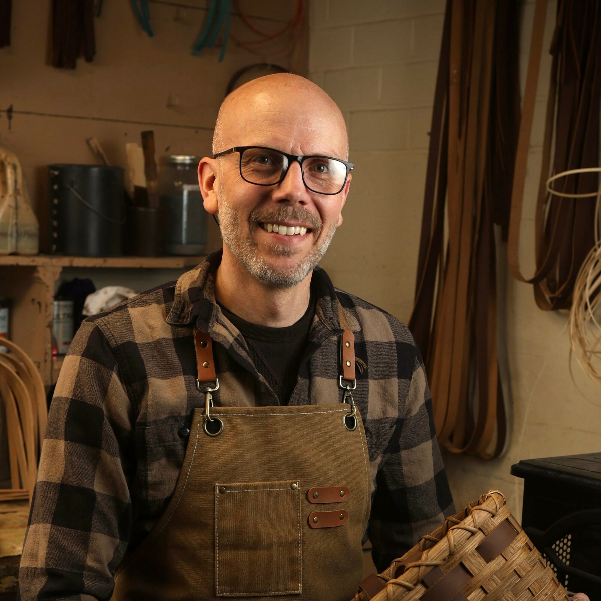 Bald man with glasses smiling, wearing a plaid shirt and apron, in a workshop or studio setting.