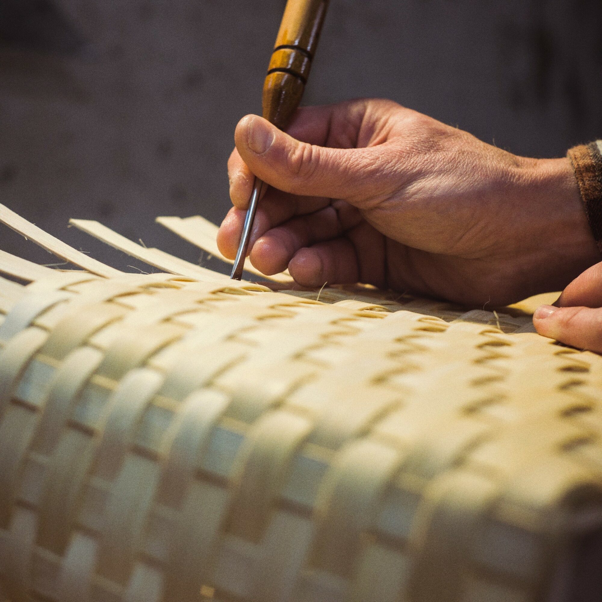 Close-up of a hand using a tool to weave or craft a woven material, with a textured background.