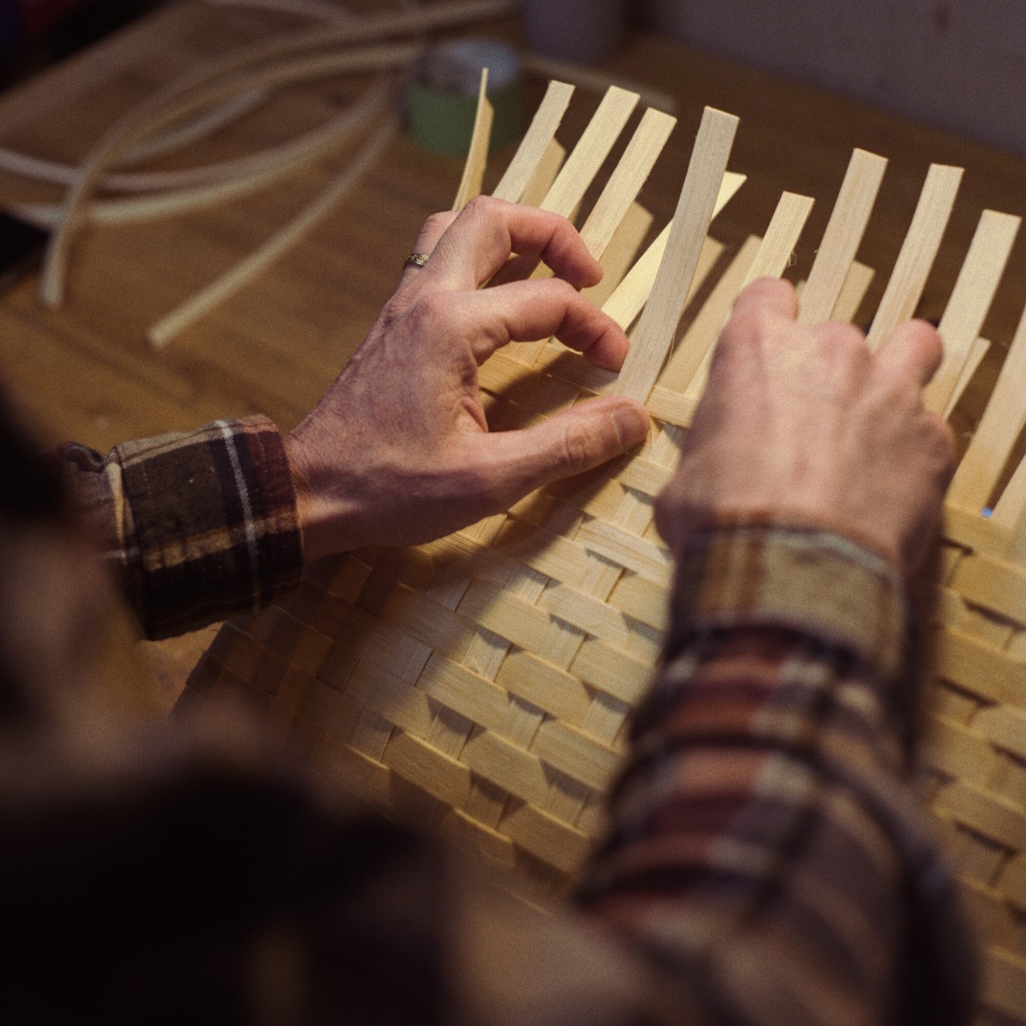 Hands of a person weaving strips of material on a wooden loom, wearing a plaid shirt.