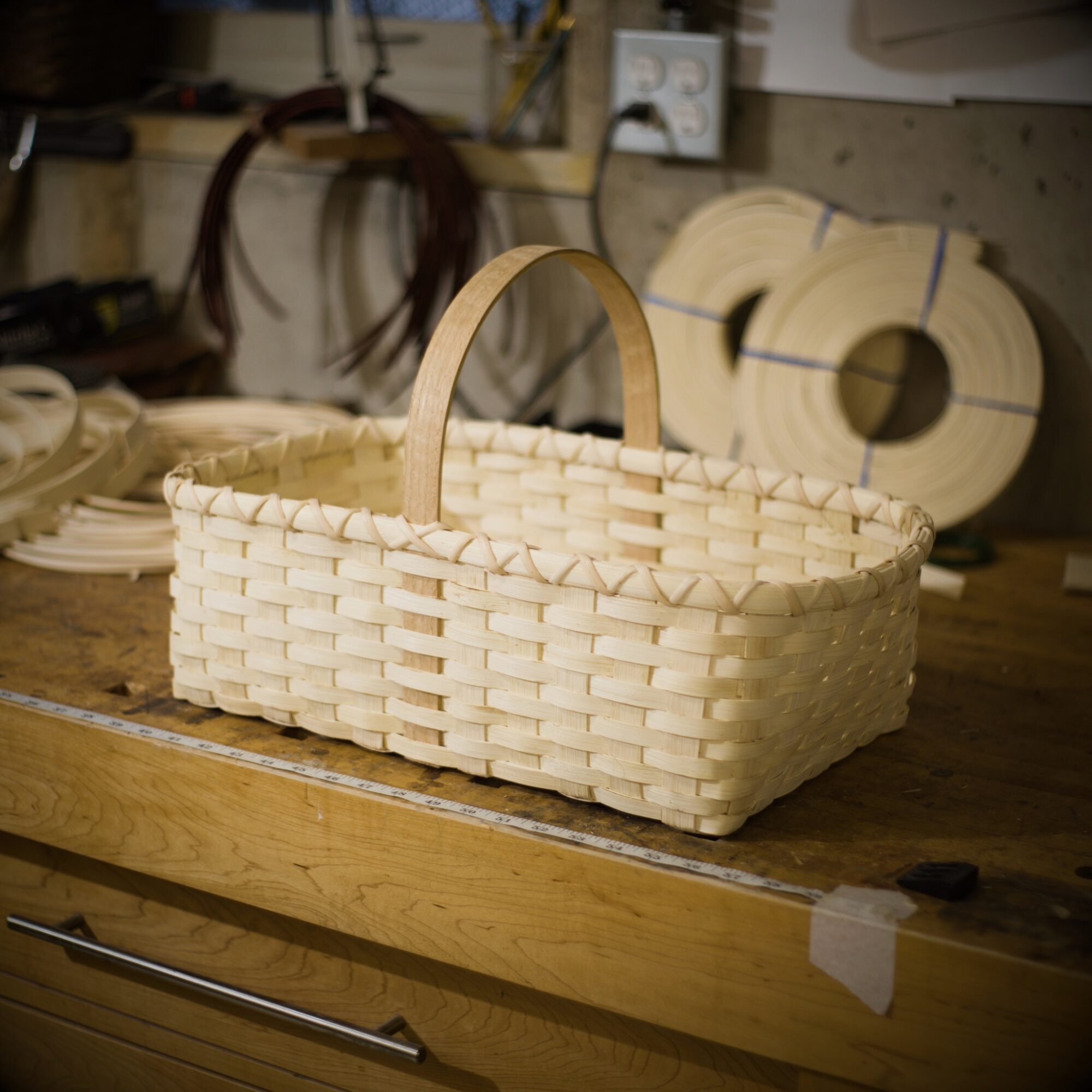 Rectangular woven basket with a handle on a wooden workbench, workshop background with tools and materials.