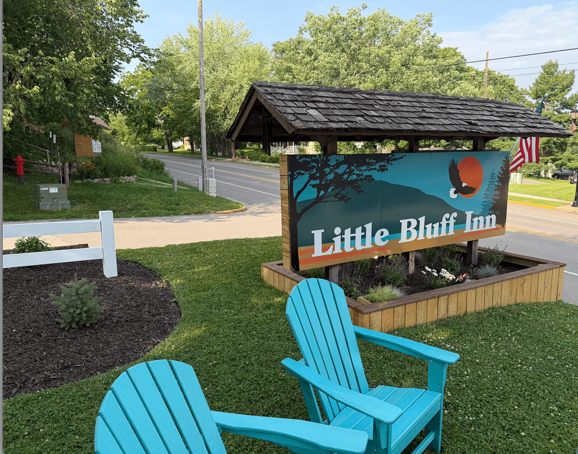 Two blue Adirondack chairs in front of a sign for Little Bluff Inn, with trees and a road in the background.