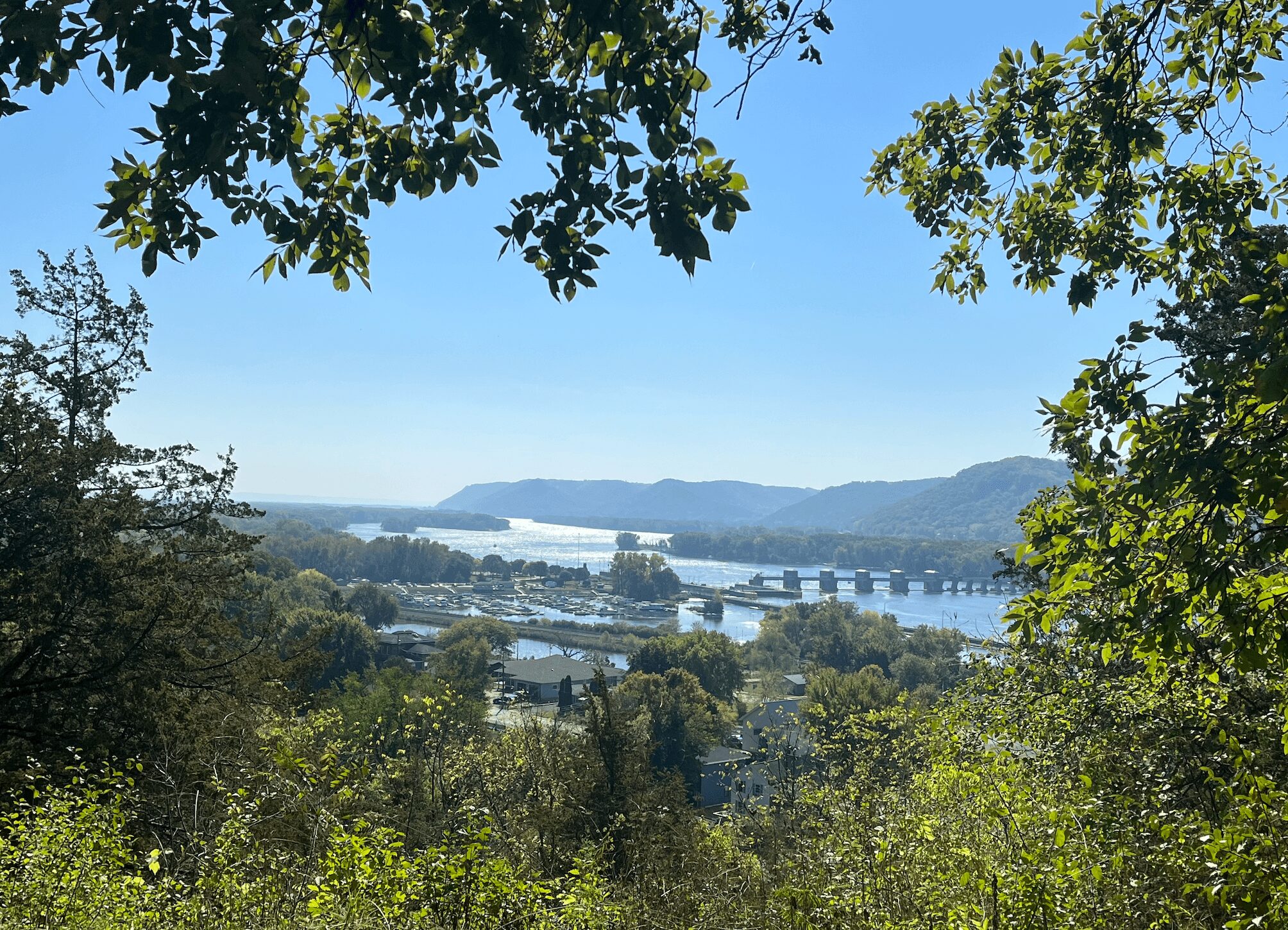View of a river with a bridge, surrounded by trees and hills in the distance, under a clear blue sky.