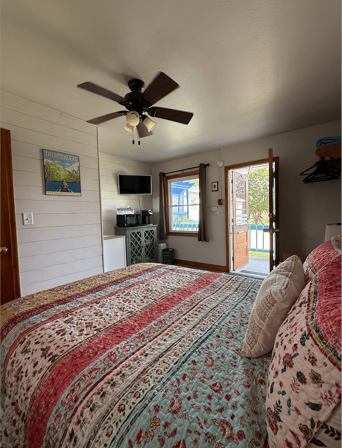 Bedroom with ceiling fan, open door to outside, bed with patterned quilt, pillows, and a small TV on a stand.