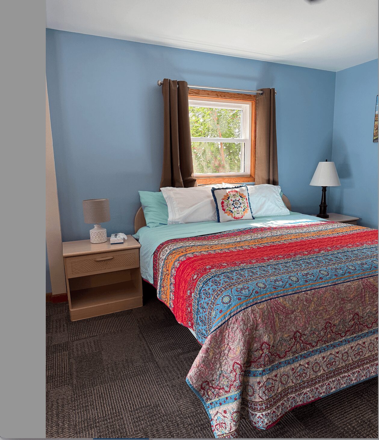 Bedroom with blue walls, window with brown curtains, bed with colorful quilt, pillows, nightstand, and lamp.