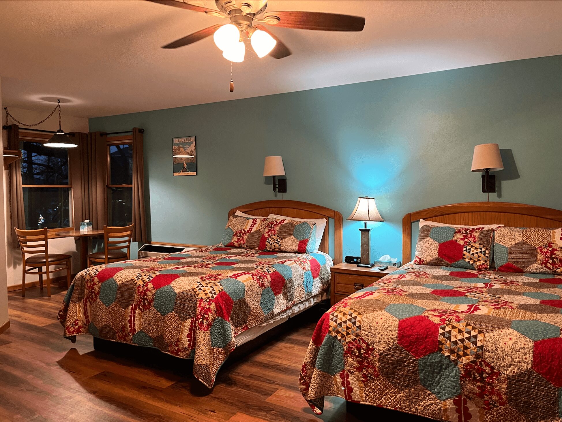 Two beds with colorful patchwork quilts in a room with teal walls, wooden furniture, and a small dining area with windows.