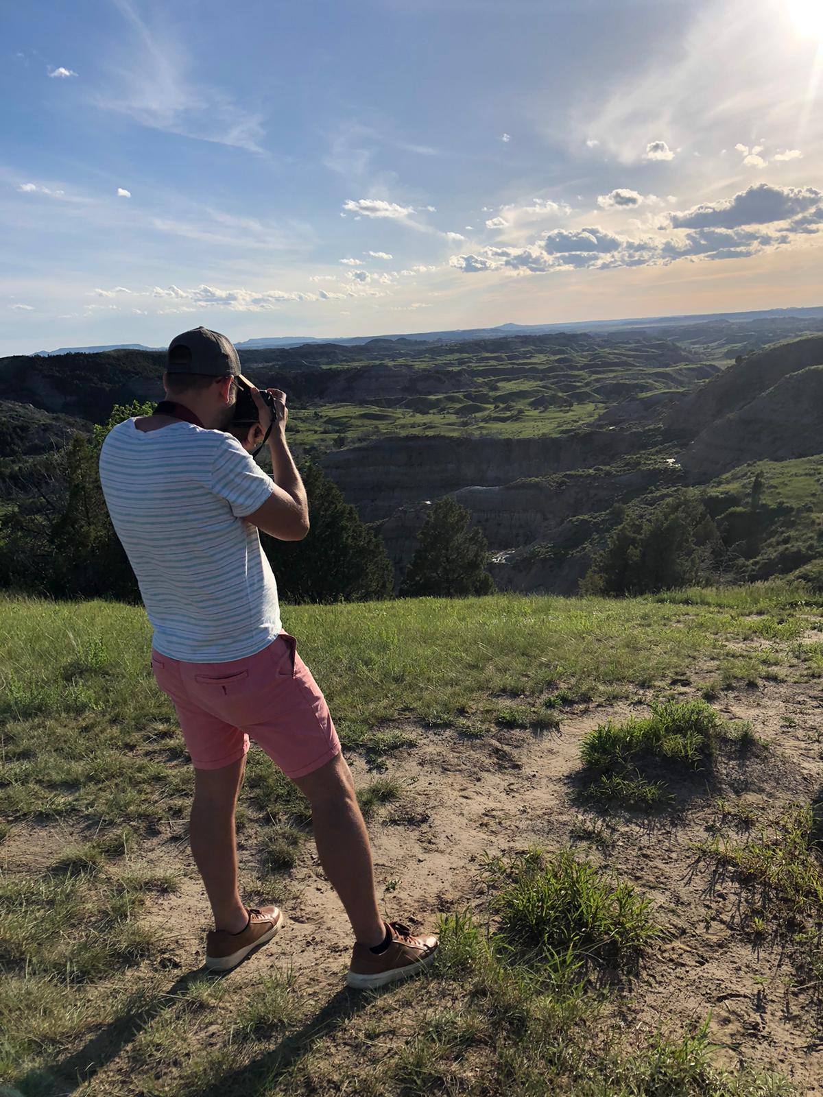 Person taking a photo outdoors on a grassy hill with a landscape and sky in the background.