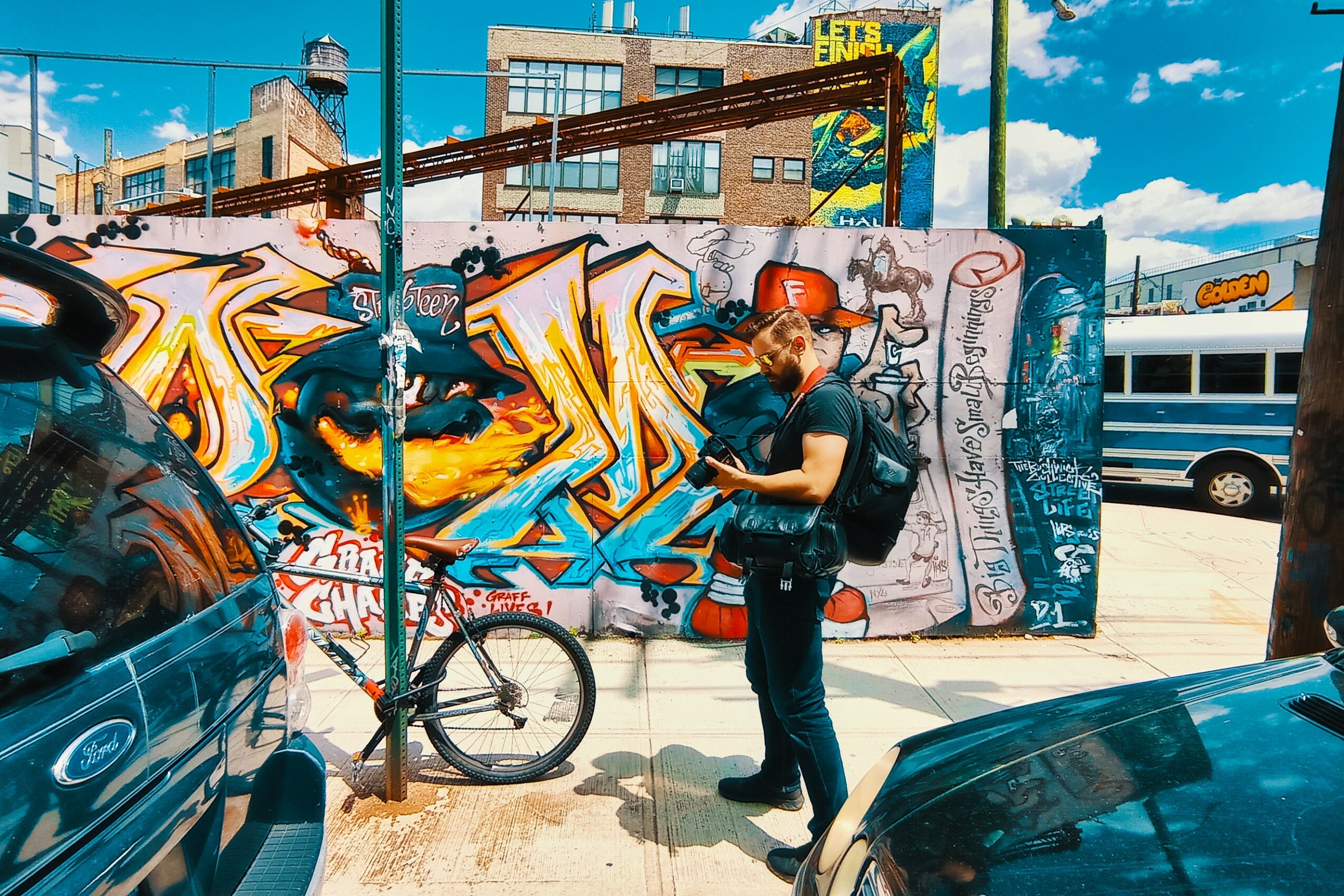 Person with a backpack standing on sidewalk, looking at phone, graffiti-covered wall behind, bicycle parked nearby, city street scene.