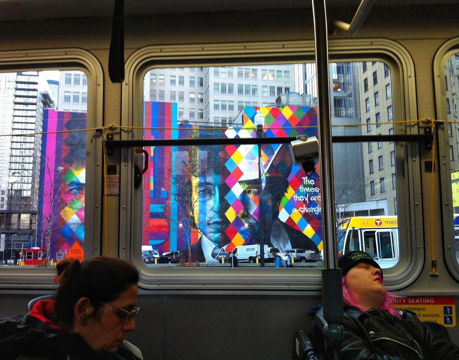 View through bus window showing colorful mural of a person's face with geometric patterns, city buildings, and a bus outside.