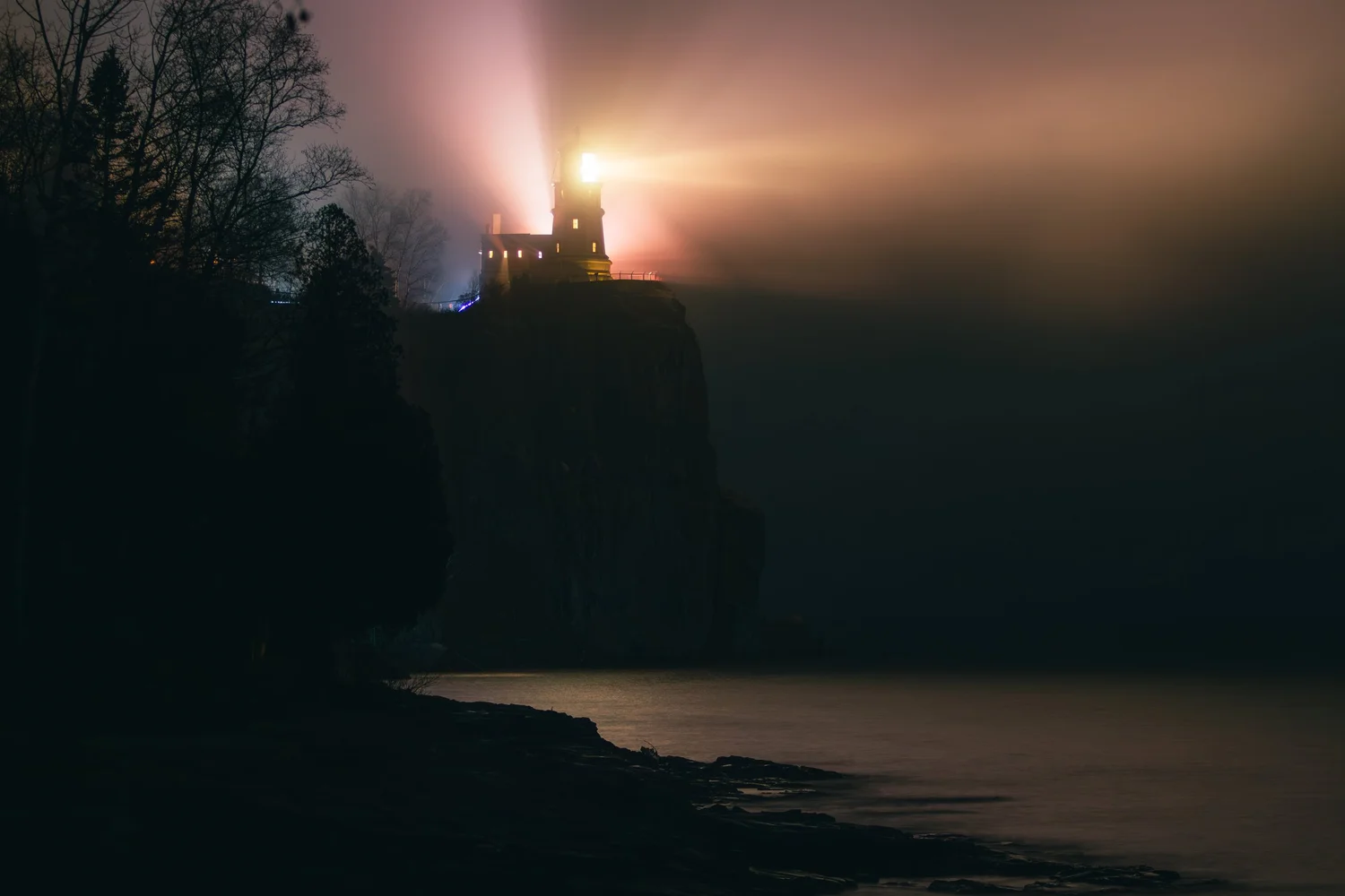 Lighthouse on a cliff emitting bright light at night, with dark trees and water in the foreground.