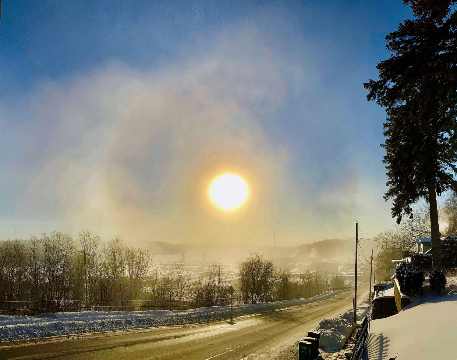 Bright sun in a clear sky over a snowy landscape with trees and a road, early morning or late afternoon light.