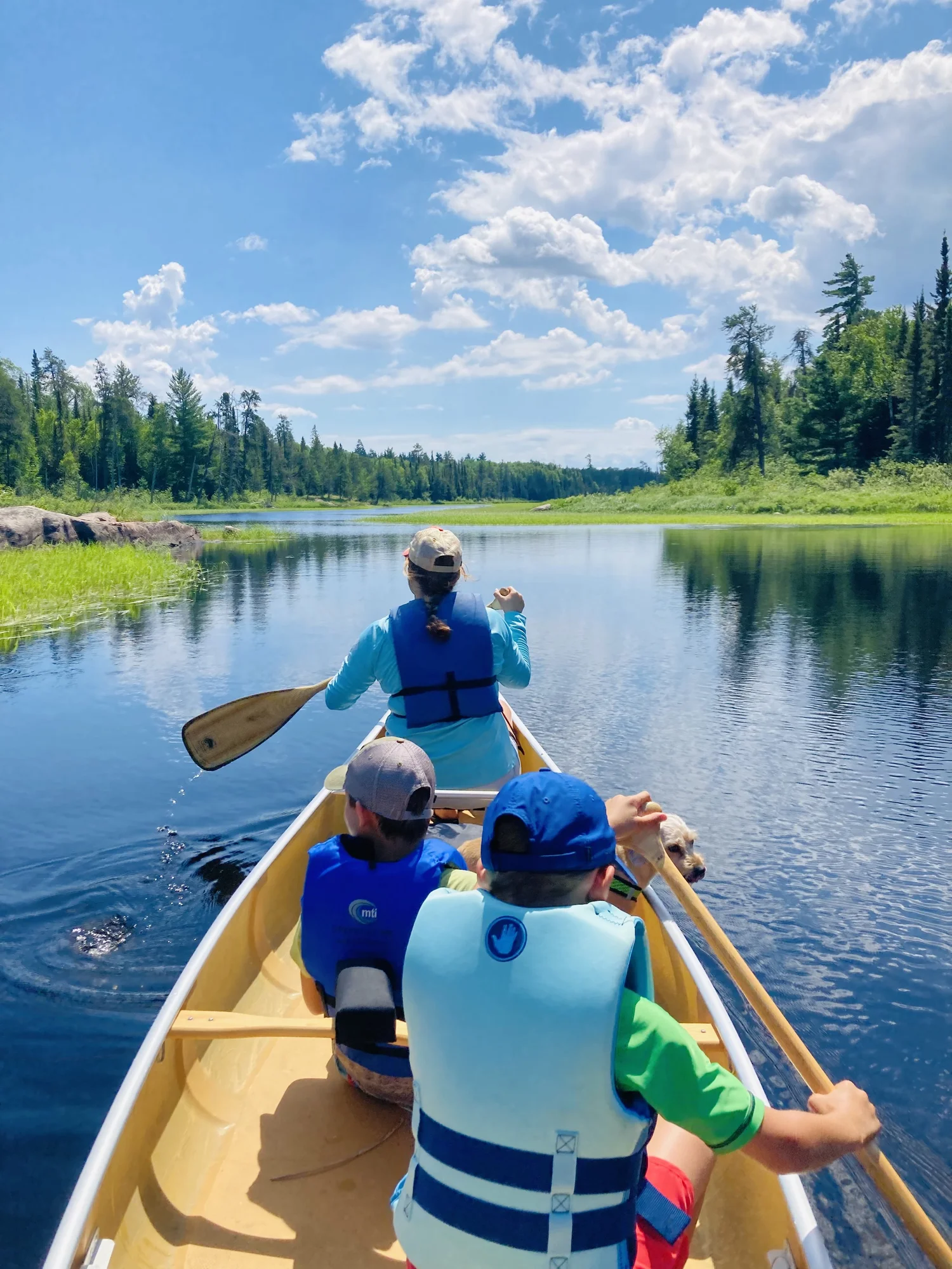 People in a yellow canoe paddling on a calm river surrounded by trees and blue sky with clouds.