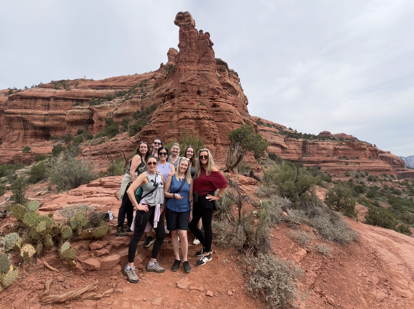 Group of seven people standing on rocky terrain with red rock formations and sparse desert vegetation in the background.