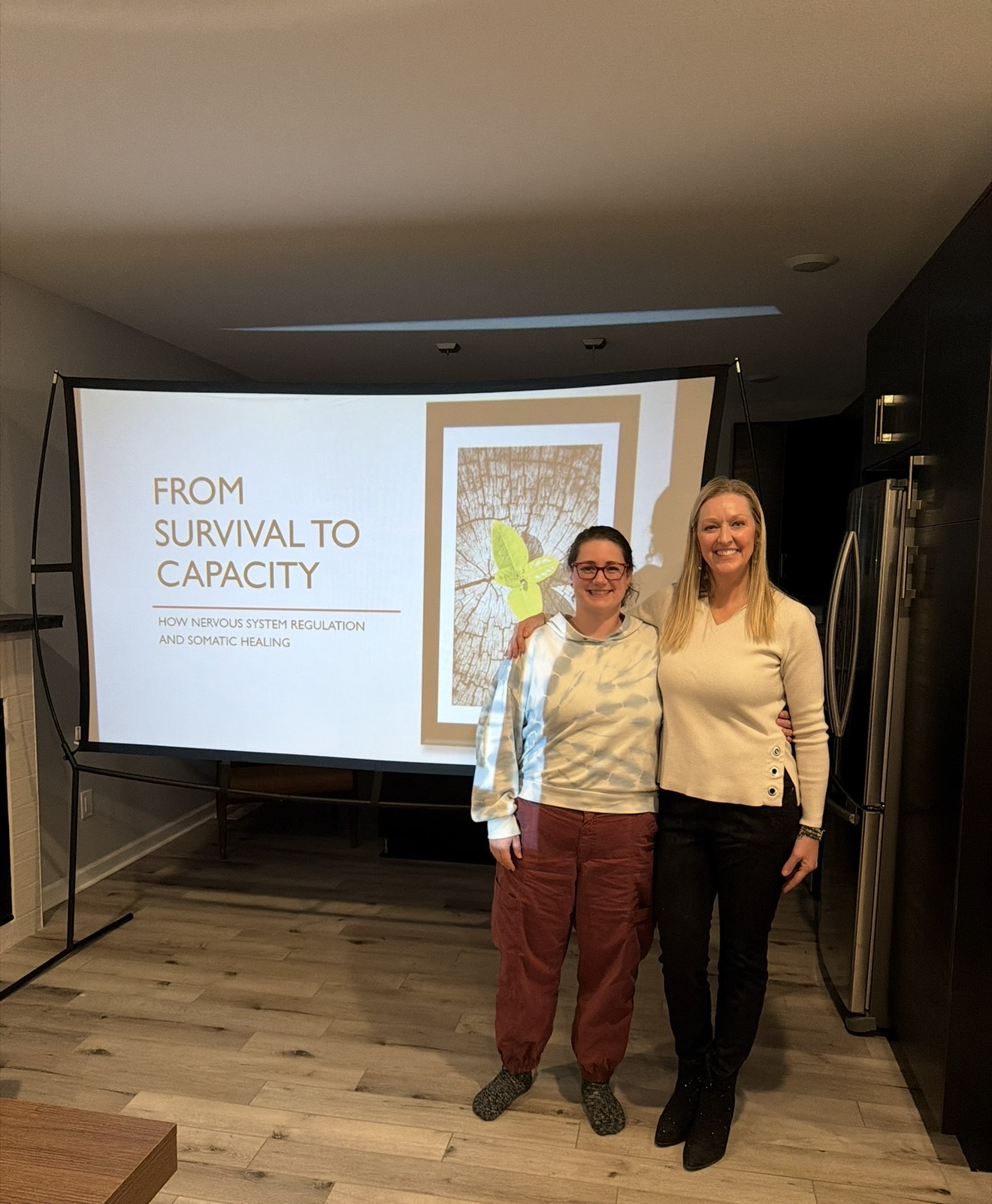 Two women stand in front of a presentation screen in a conference room, smiling at the camera.