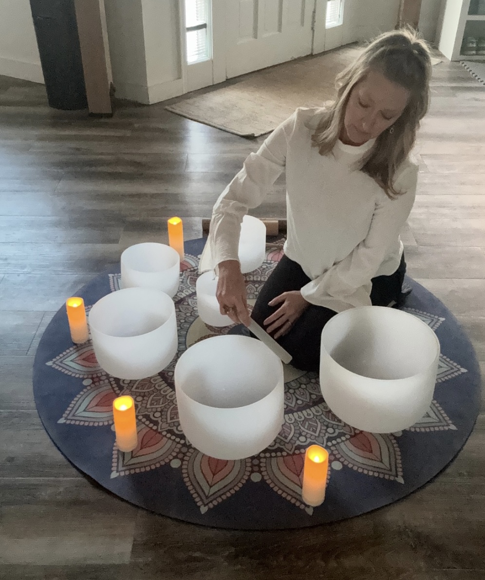 Girl sitting on a round rug with white singing bowls and orange candles inside a home, wooden floor and door in background.