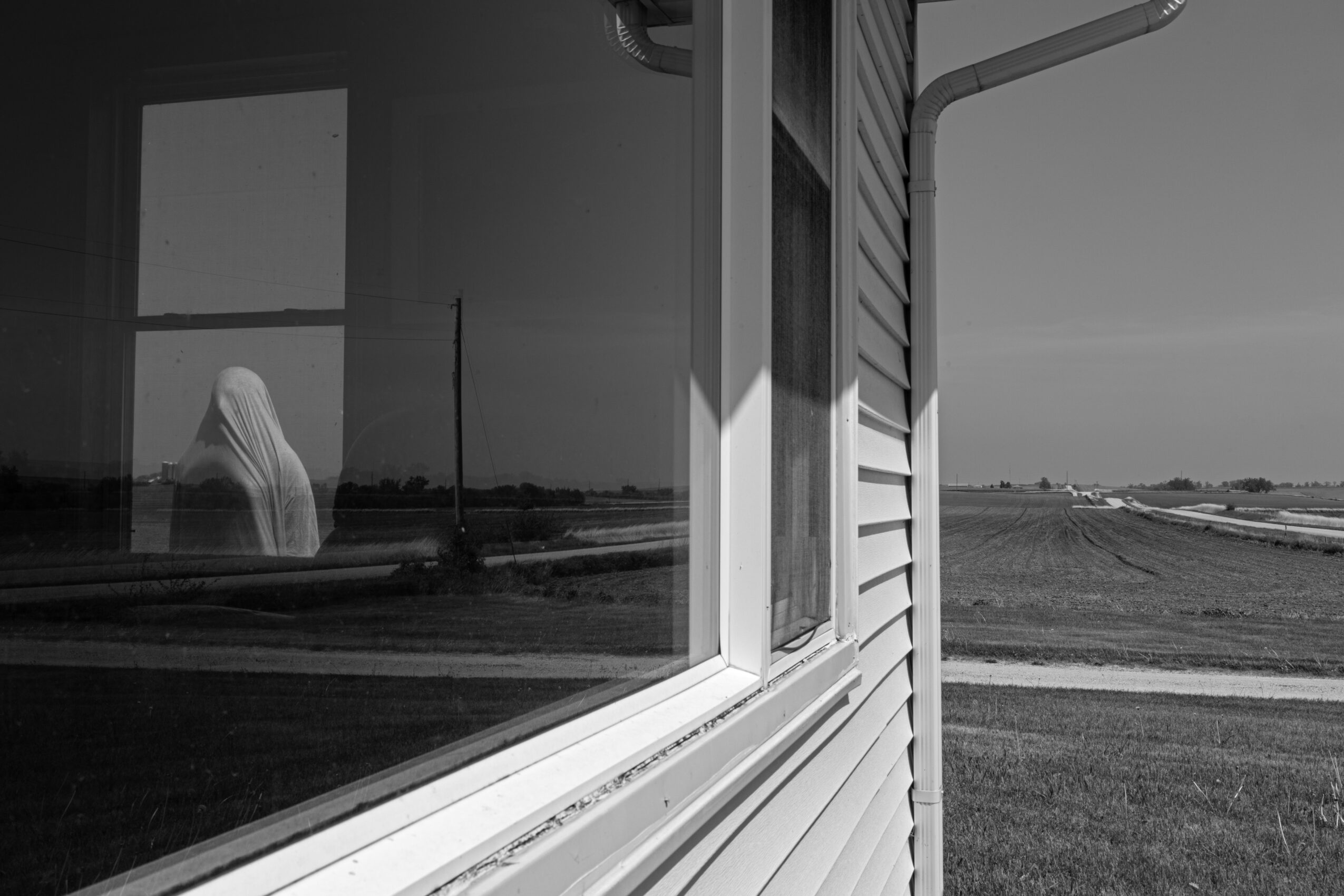 View of a window on the side of a house with a rural landscape outside, including fields and a dirt road.