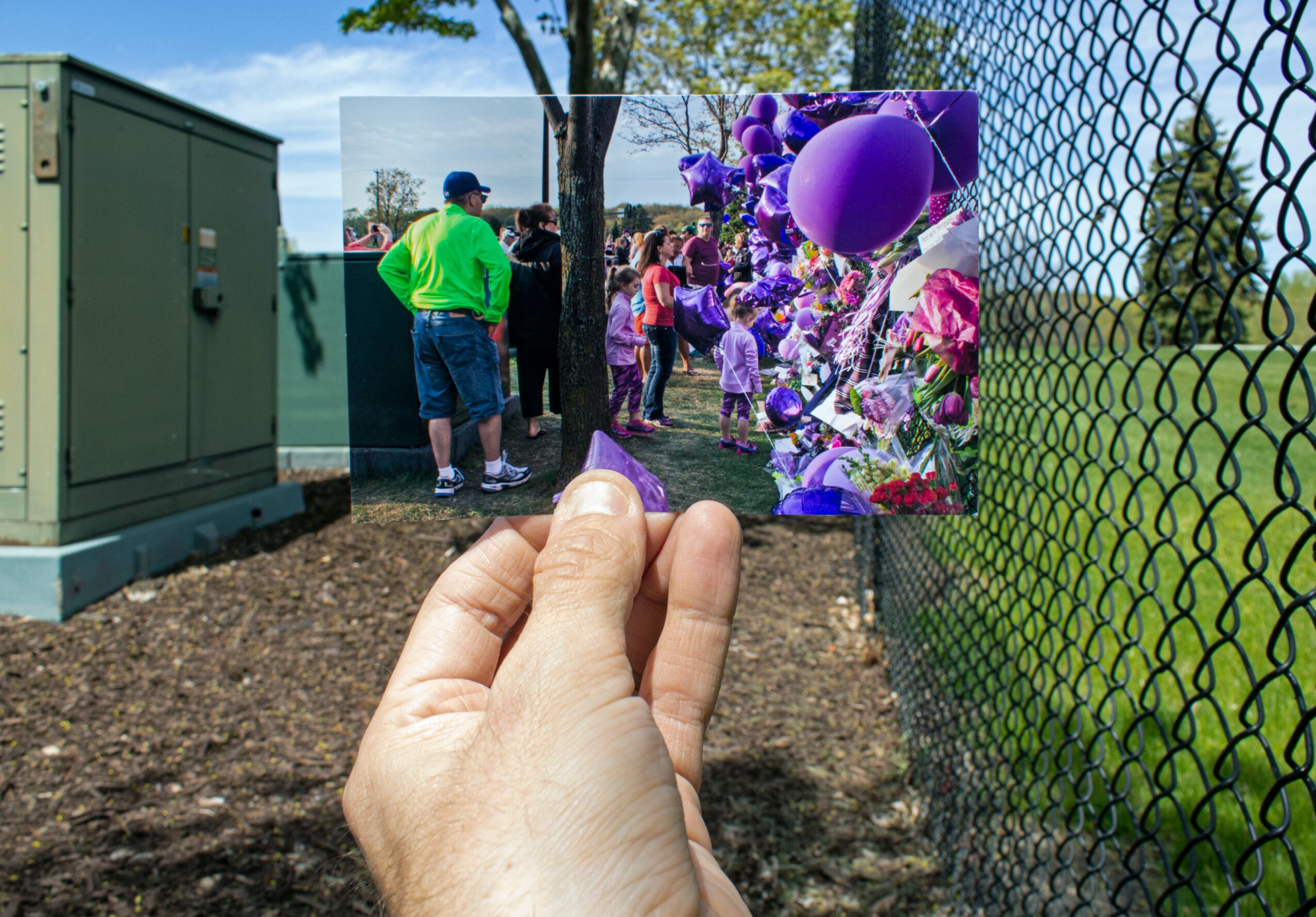 Person holding a photo of children and balloons outdoors, with a fence and trees in the background.