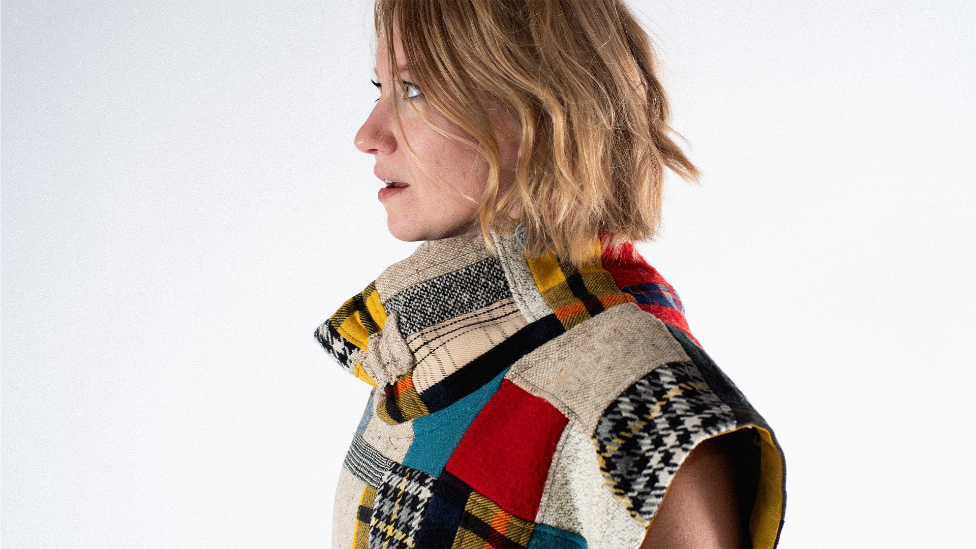 Side profile of a woman with short, wavy hair wearing a colorful plaid scarf and patterned coat against a white background.
