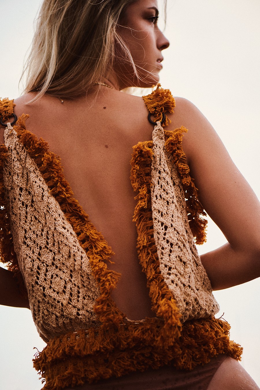 Side view of a woman wearing a crochet top with fringes, showing her back and shoulder, against a light background.