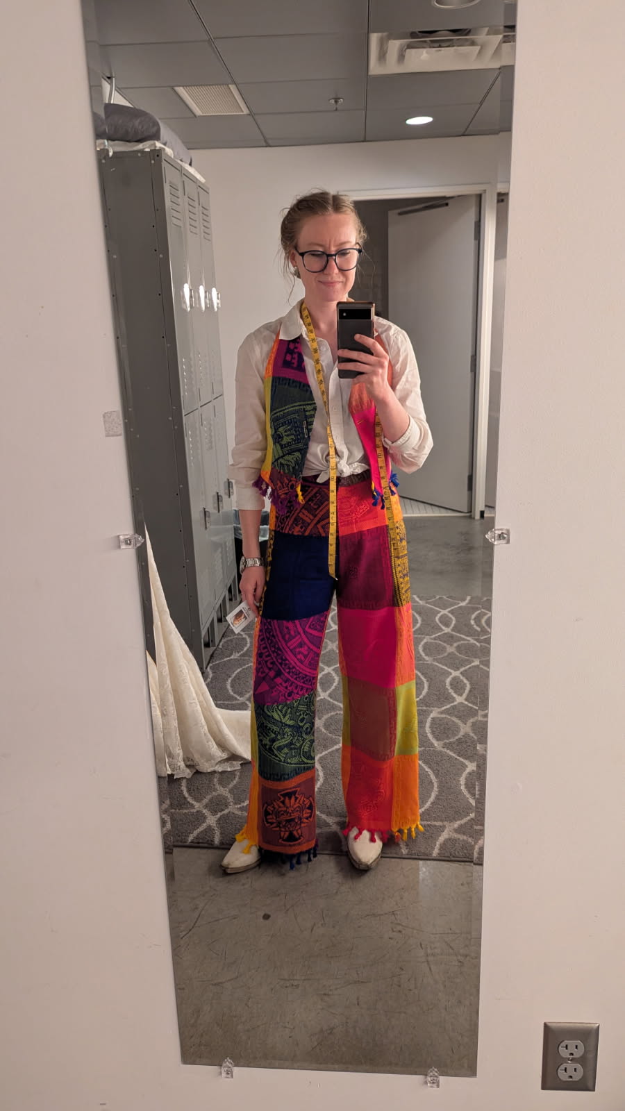 Person taking a mirror selfie in a room, wearing colorful patterned pants and a light-colored jacket, holding a phone.