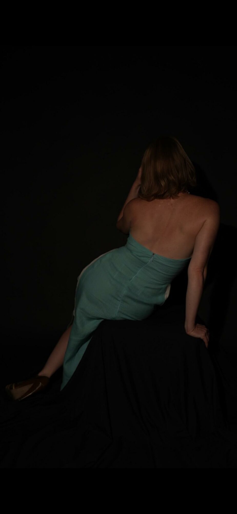 Person with long hair, back facing, sitting on a black surface, wearing a strapless dress, against a dark background.