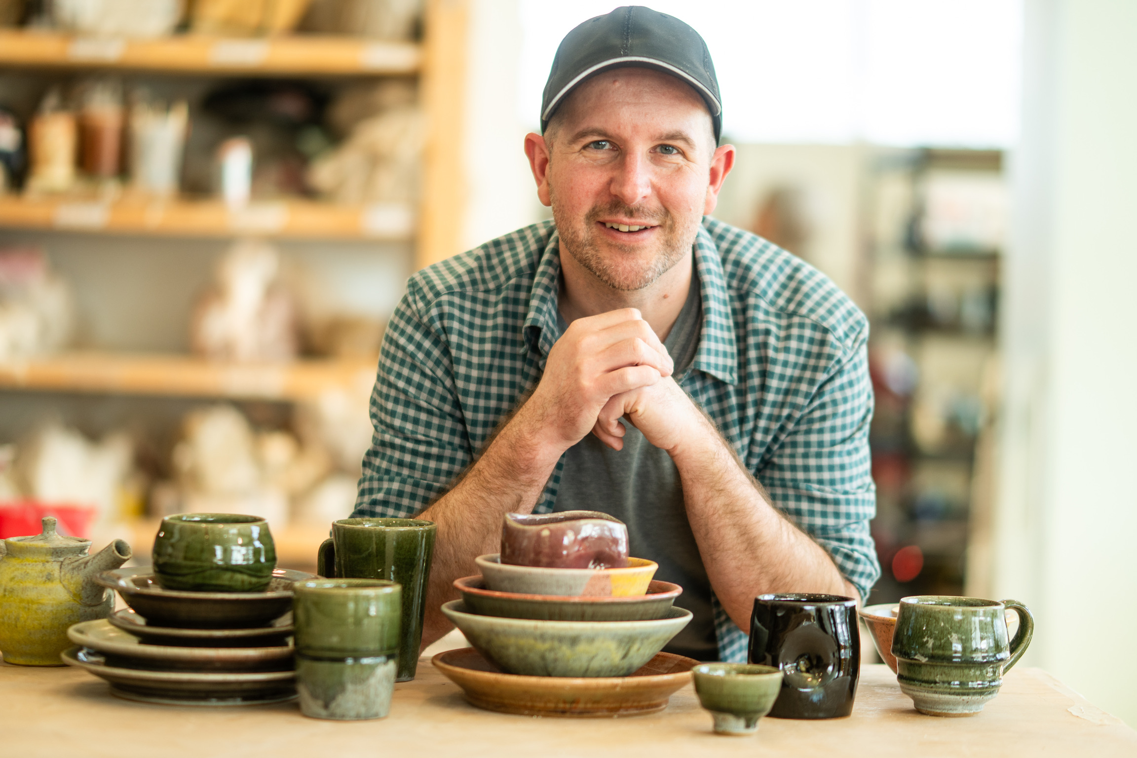 Man with a beard and cap smiling, sitting at a table with stacked bowls and cups, in a bright room.