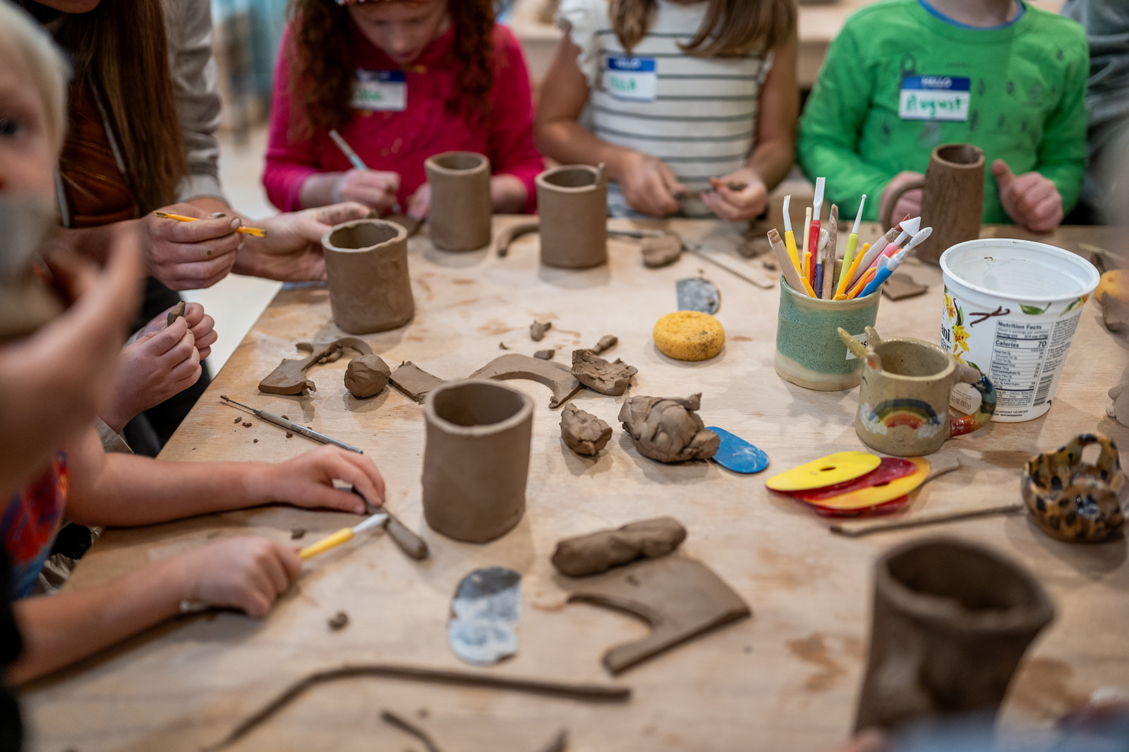 Children gather around a table with clay, tools, and art supplies, working on creative projects together.