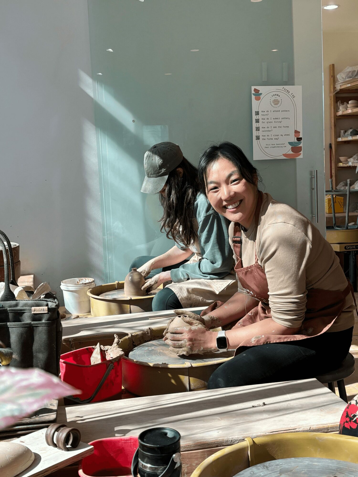 Woman smiling while washing dishes at a sink with a person in the background, sunlight streaming in, kitchen setting.
