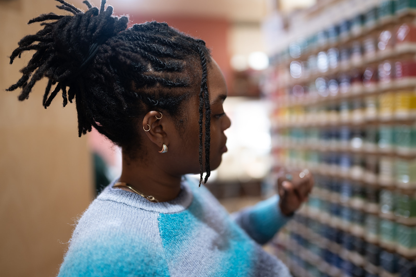 Woman with dreadlocks looking at shelves of colorful products in a store, wearing a light blue sweater, side profile.