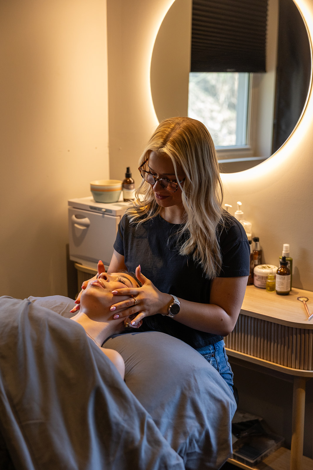 A woman with glasses and blonde hair gives a facial treatment to a person lying on a bed in a spa room with a round illuminated mirror.