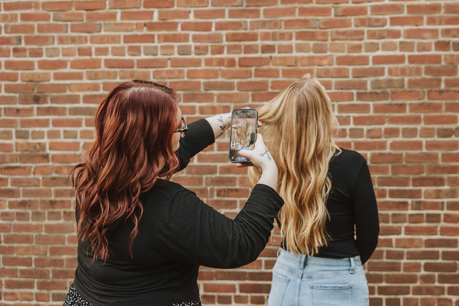 Two women with long hair, one with red and the other with blonde, stand against a brick wall, taking a photo.