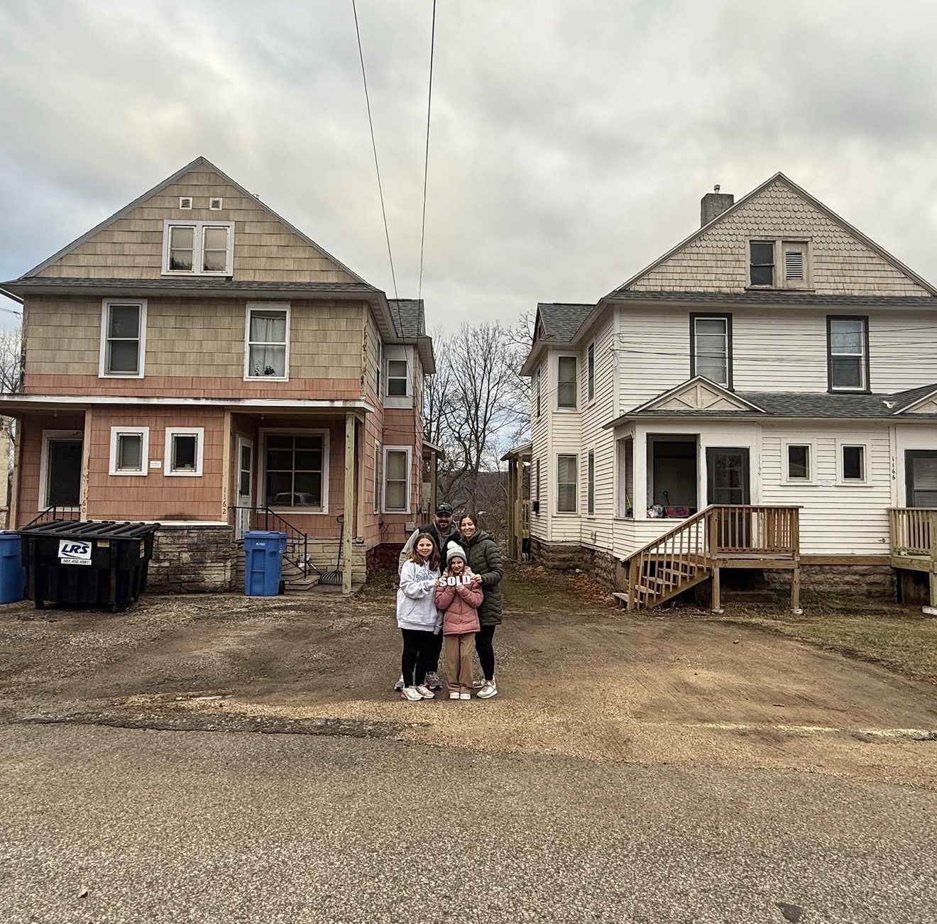 Three people standing on a dirt road in front of two houses, with cloudy sky overhead.