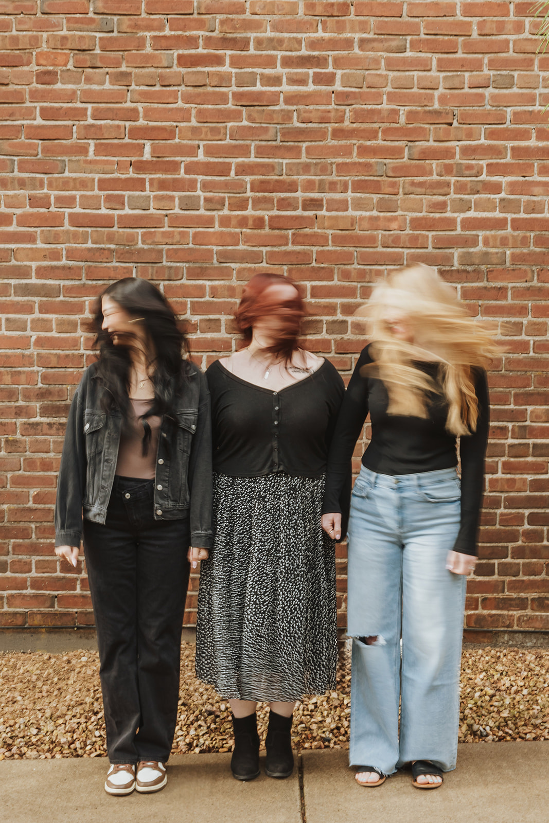 Three women standing against a brick wall, hair blowing, wearing casual clothing, with a gravel and concrete ground.