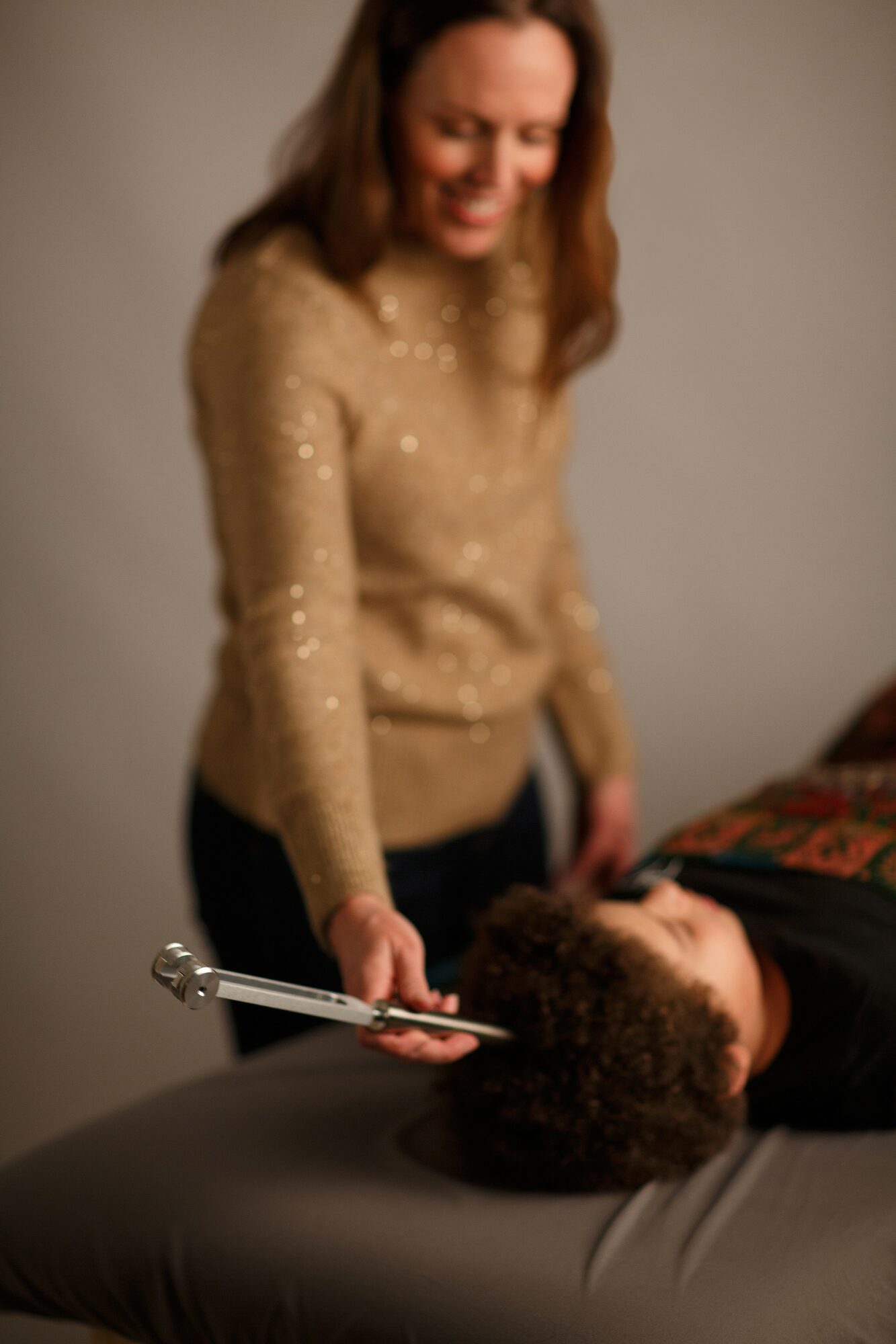 Woman smiling and holding a metal tool over a person lying on a table, in a neutral room.
