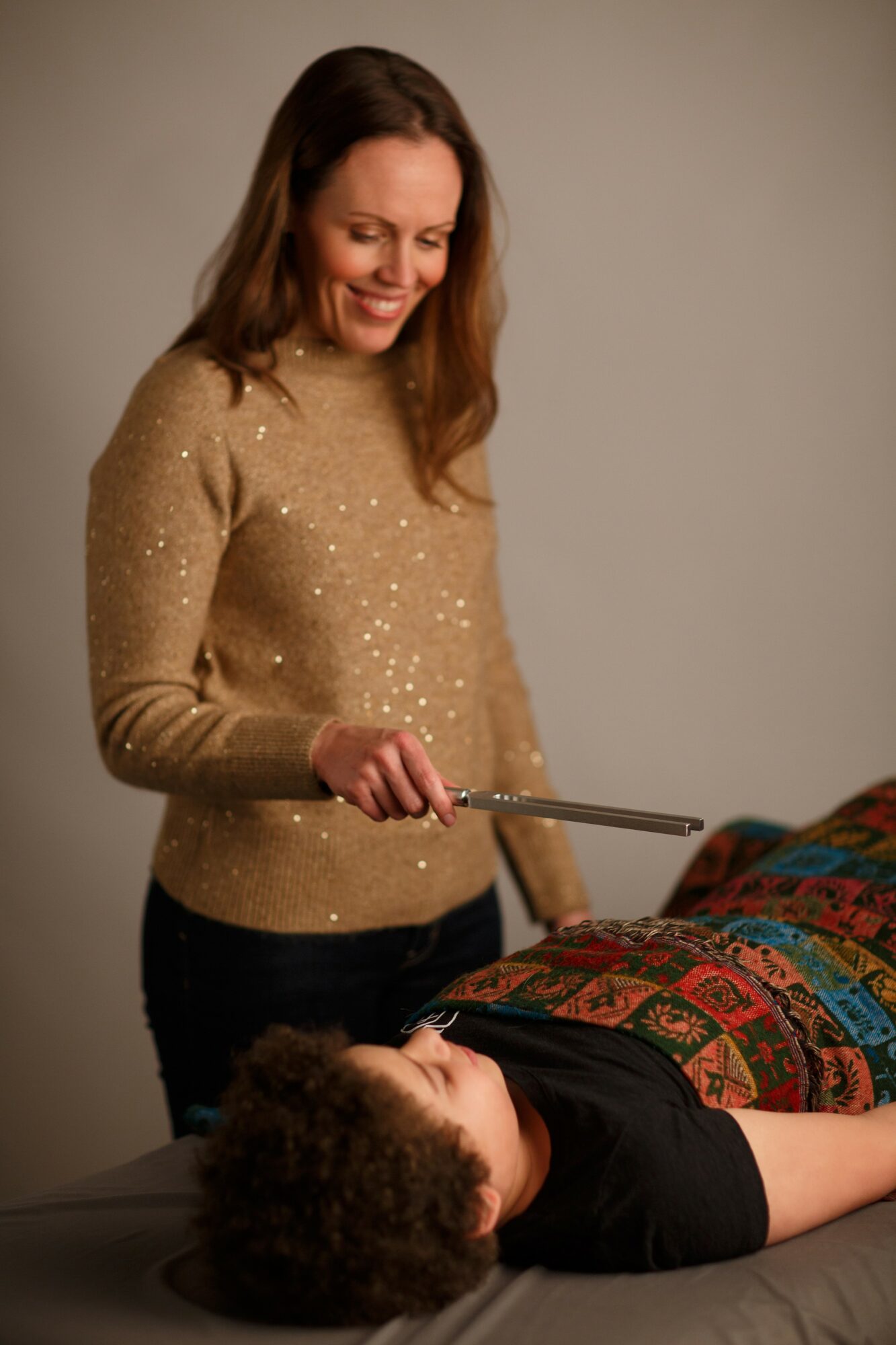 Woman smiling and holding a stick over a person lying on a table, with colorful fabric nearby.