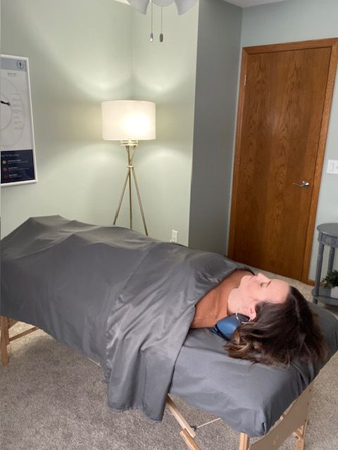 Person lying on a gray massage table in a room with a wooden door and a floor lamp.