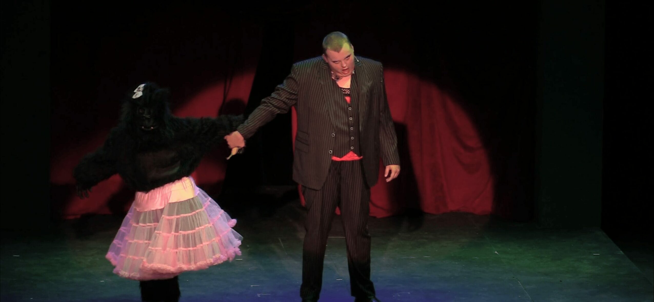 Two performers on stage, one in a dark suit and the other in a colorful skirt, holding hands.