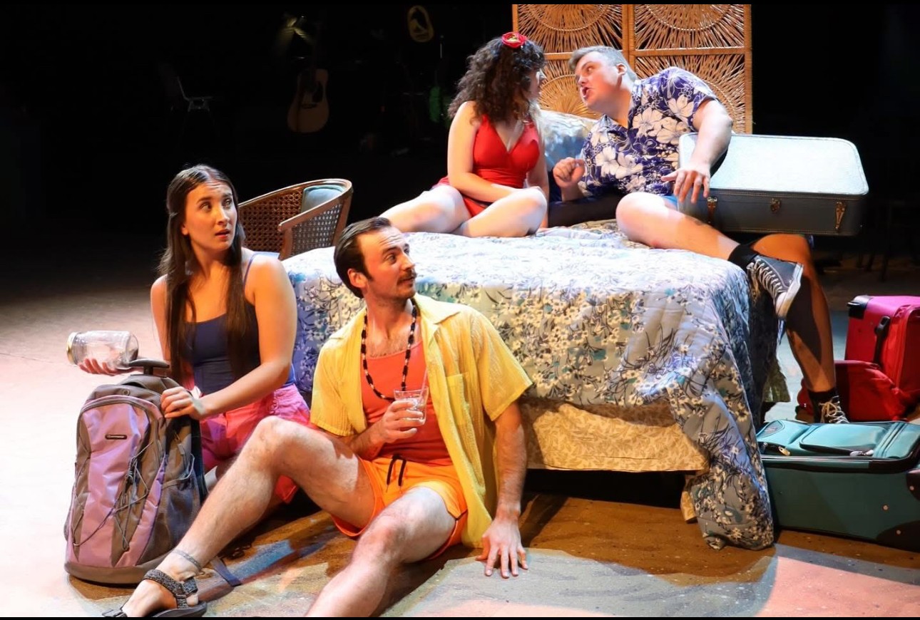 Four people on a stage with a bed, two sitting on it and two sitting on the floor, with luggage nearby.