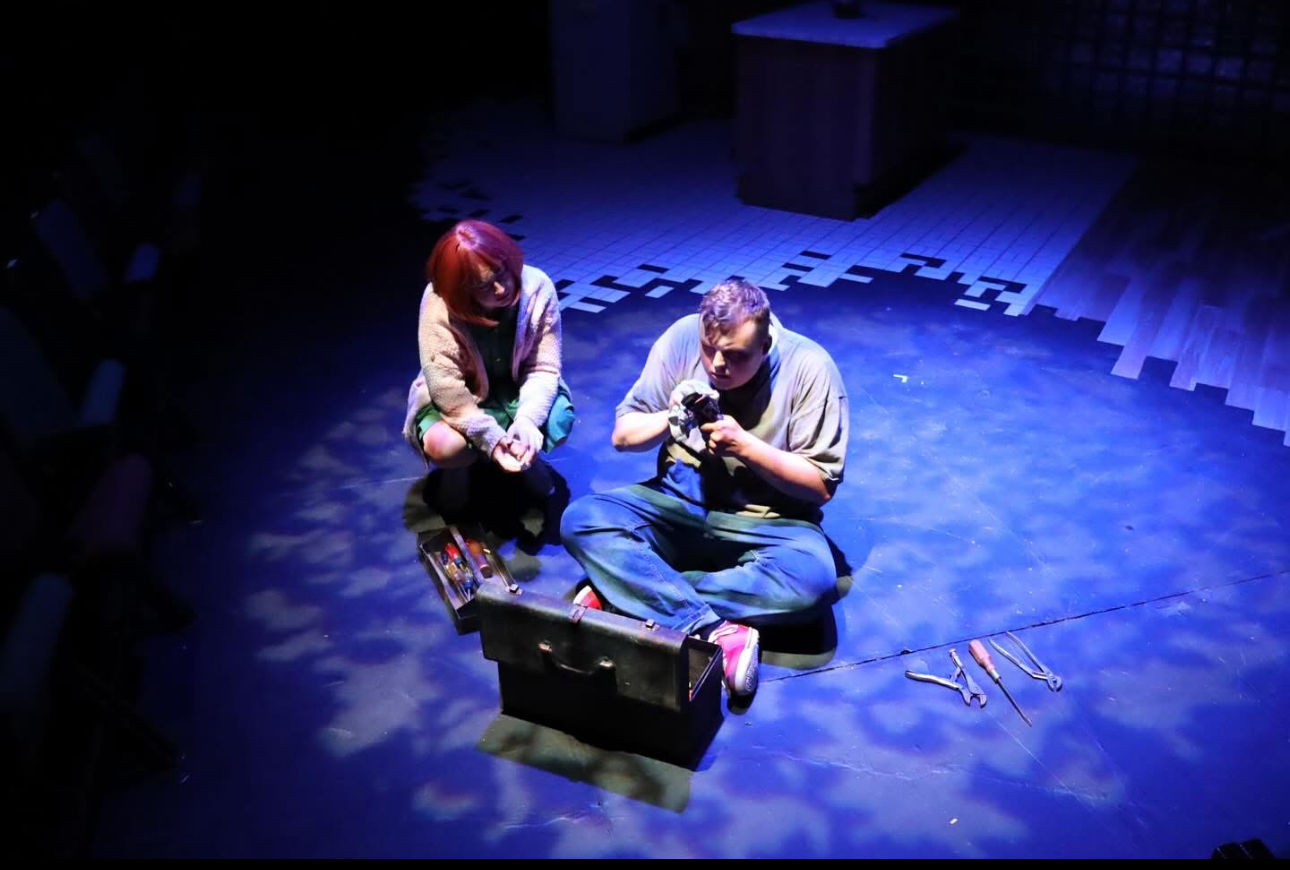Two people sit on the floor in a dark room with blue lighting, surrounded by a box and tools, engaging in an activity.